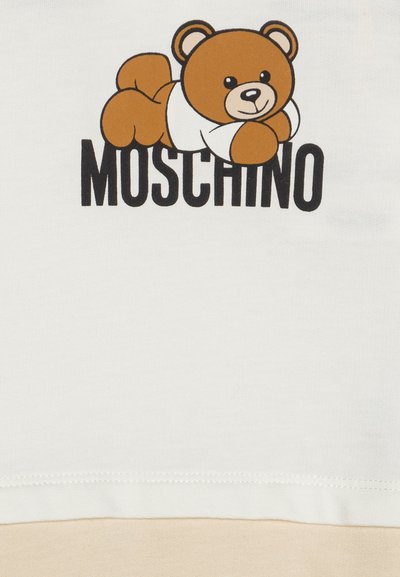 T-shirt di cotone color crema con grafica di un orso marrone in stile cartoon e testo nero "Moschino" sotto. Tessuto morbido, design casual.