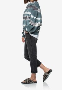 Sudadera de gran tamaño con diseño de tie-dye en teal y blanco, combinada con jeans negros desgastados y sandalias tipo slide en houndstooth negro y marrón.