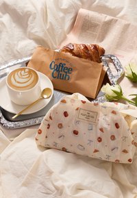 Tazza di caffè con latte art, cucchiaio d'oro, croissant in sacchetto di carta, involucro riutilizzabile decorato con illustrazioni di cibo, vassoio d'argento, sfondo in tessuto bianco.