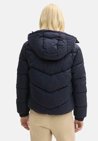 Námornícky modrá nadýchaná bunda s kapucňou, ktorá má vodorovné a chevronové švy, elastické manžety a skrátený strih.