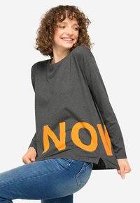 Maglietta grigia a maniche lunghe con la parola "NOW" in grandi lettere arancioni nella parte inferiore. Il materiale sembra morbido con una vestibilità rilassata e fenditure laterali.