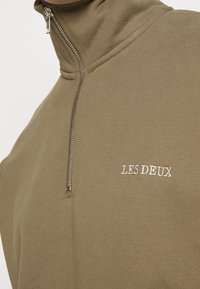 Sweat-shirt zippé vert olive avec un col montant, présentant un tissu texturé subtil et un logo brodé "LES DEUX" en blanc.