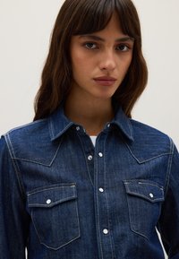 Young woman with long brown hair wearing a dark blue denim button-up shirt with two front flap pockets.