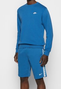 Ensemble Nike bleu composé d'un sweat-shirt et d'un short, fabriqué en tissu doux. Il présente un col rond, un logo brodé et une bande latérale contrastée sur le short.