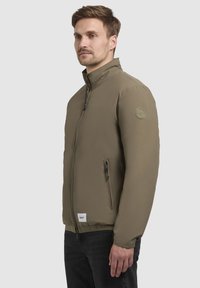 Oliven grøn zip-up jakke med høj krave, frontlommer og et cirkulært logoemblem på venstre skulder. Glat tekstur, minimalistisk design.