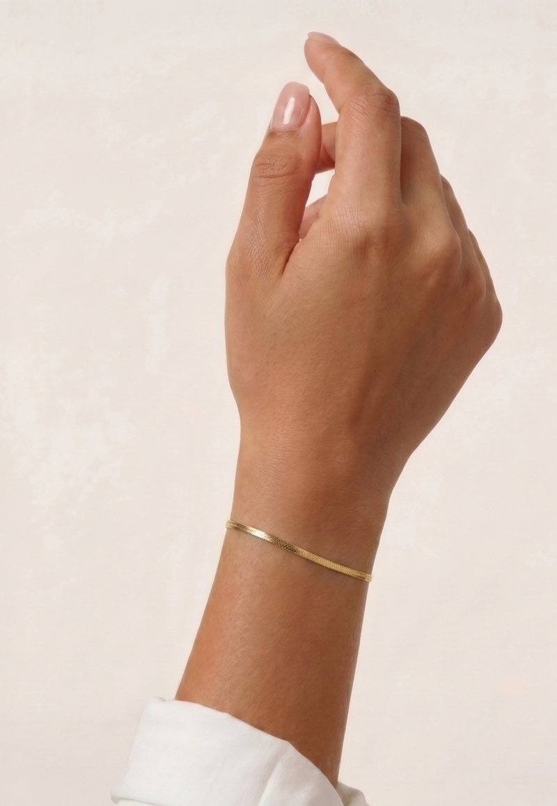 Main droite avec un fin bracelet en or au poignet sur un fond clair uni, doigts légèrement recourbés.