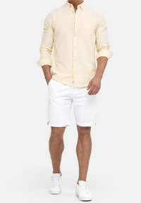 Lichtgele linnen shirt met opgerolde mouwen, gecombineerd met witte katoenen shorts. Model draagt witte sneakers, eenvoudig ontwerp met een voorzak.