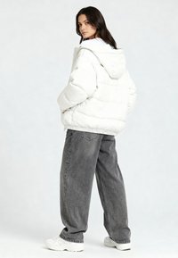 Giacca imbottita bianca con cappuccio, caratterizzata da un design trapuntato, orlo elastico e due tasche laterali, abbinata a jeans grigi larghi e sneakers bianche.