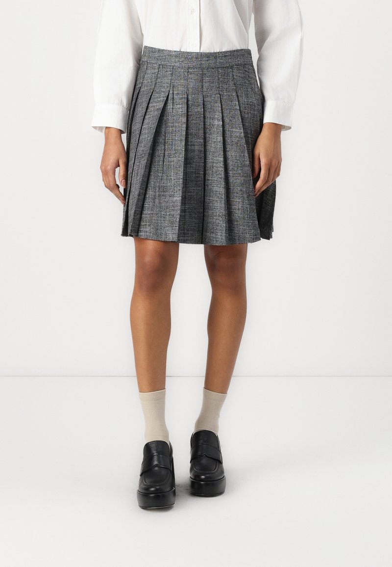Femme portant une jupe plissée grise, une chemise blanche boutonnée, des chaussettes beiges et des mocassins noirs, debout devant un fond blanc uni.