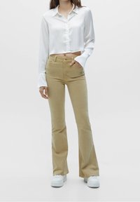 Chemise blanche courte à boutons avec manches longues, associée à un pantalon beige évasé et des baskets blanches à plateforme. Tissu lisse, taille cintrée.