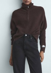Rjav rebrast turtleneck pulover z zadrgo do polovice. V kombinaciji z črnimi kavbojkami visokega pasu in srebrnimi manšetami na rokavih.