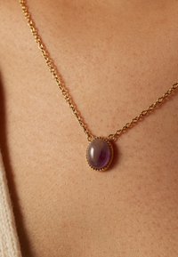 Gouden ketting met een ovale, paarse edelsteenpendant met een glad oppervlak en decoratieve kraalaccenten langs de rand.