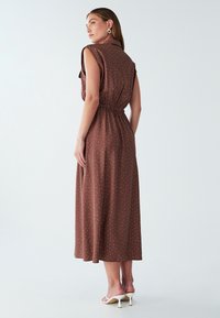 Brązowa sukienka midi w białe kropki, krótki rękaw, marszczona talia i gładka tekstura. Stylizowana z białymi sandałami na wysokim obcasie.