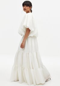 Weißes, strukturiertes langes Kleid mit einem puffigen, kurzärmeligen Oberteil und schichtbarem Rock. Das Design verfügt über eine geraffte Taille und eine fließende Silhouette.