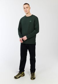 Groene langesleeve T-shirt met een klein logo op de borst, gecombineerd met donkere spijkerbroeken en schoenen met camouflagepatroon. Eenvoudig ontwerp, casual pasvorm.