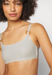 Bralette cinza claro com tecido texturizado, design simples e detalhes decorativos em madeira nas alças ajustáveis.