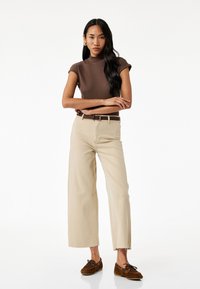 Bruine geribbelde coltrui, beige wijde broek met rafelende zoom en bruine suède instapschoenen, met een bijpassende riem.