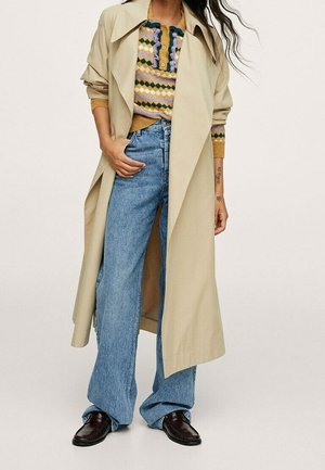 Beige-Trenchcoat über einem gemusterten Pullover mit Knopfleiste, kombiniert mit weiten blauen Jeans und dunklen Lederschuhen.
