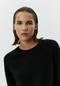 Pulover negru cu guler rotund, cu o textură moale, având o croială lejeră și mâneci integrate. Modelul are părul drept și machiaj natural.