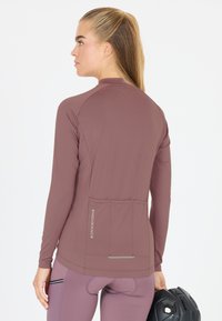 Vrouw die een mauve lange mouw fietsshirt en legging draagt, een zwarte fietshelm vasthoudend, van achteren gezien tegen een witte achtergrond.