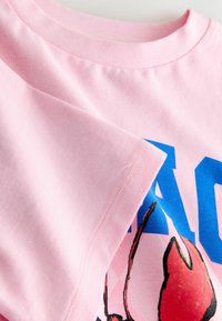 Detail růžového rukávu a výstřihu trička s modrým textem a grafikou červené klepety humra na látce.