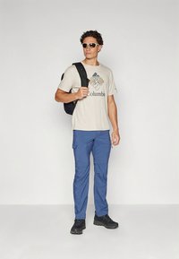 Camiseta beige para hombre con logo, pantalones cargo azules, mochila negra y zapatos negros. Modelo con gafas de sol, posando frente a un fondo liso.