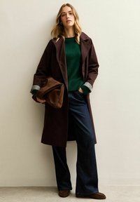 Cappotto lungo marrone con ampi risvolti, maglione verde e jeans scuri svasati. Il modello tiene una borsa marrone e indossa scarpe marroni con texture.
