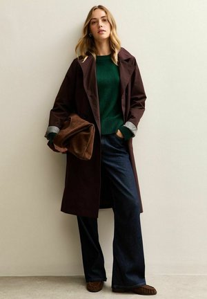Bruine trenchcoat met brede revers, groene trui en donkere flared jeans. Het model houdt een bruine tas vast en draagt gestructureerde, bruine schoenen.
