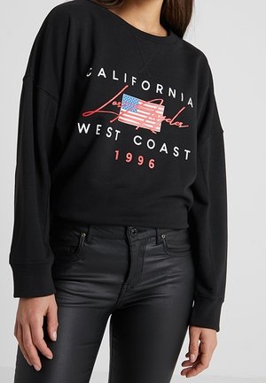 Černá mikina s nápisem "California Los Angeles West Coast 1996" a stylizovanou grafikou americké vlajky, nošená s černými kalhotami.