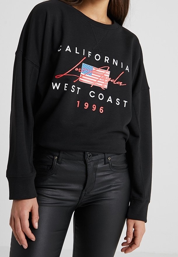 Sweat-shirt noir avec le texte "California Los Angeles West Coast 1996" et un graphisme stylisé du drapeau américain, porté avec un pantalon noir.