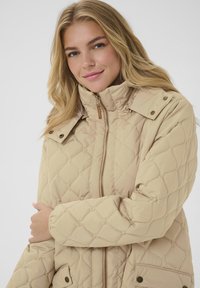 Veste matelassée beige avec un col montant, fermeture zippée et accents à boutons-pression. Elle présente une texture douce et un motif en losanges sur l'ensemble.