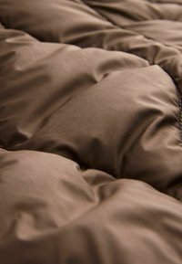 Braune Bettdecke mit glatter, matter Oberfläche und gesteppter Struktur, die ein weiches, gepolstertes Aussehen erzeugt.