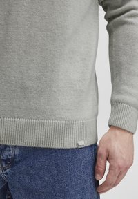 Pull en tricot gris clair avec des manches et un ourlet côtelés, présentant une texture subtile et une petite étiquette de marque sur le côté, associé à un jean bleu.