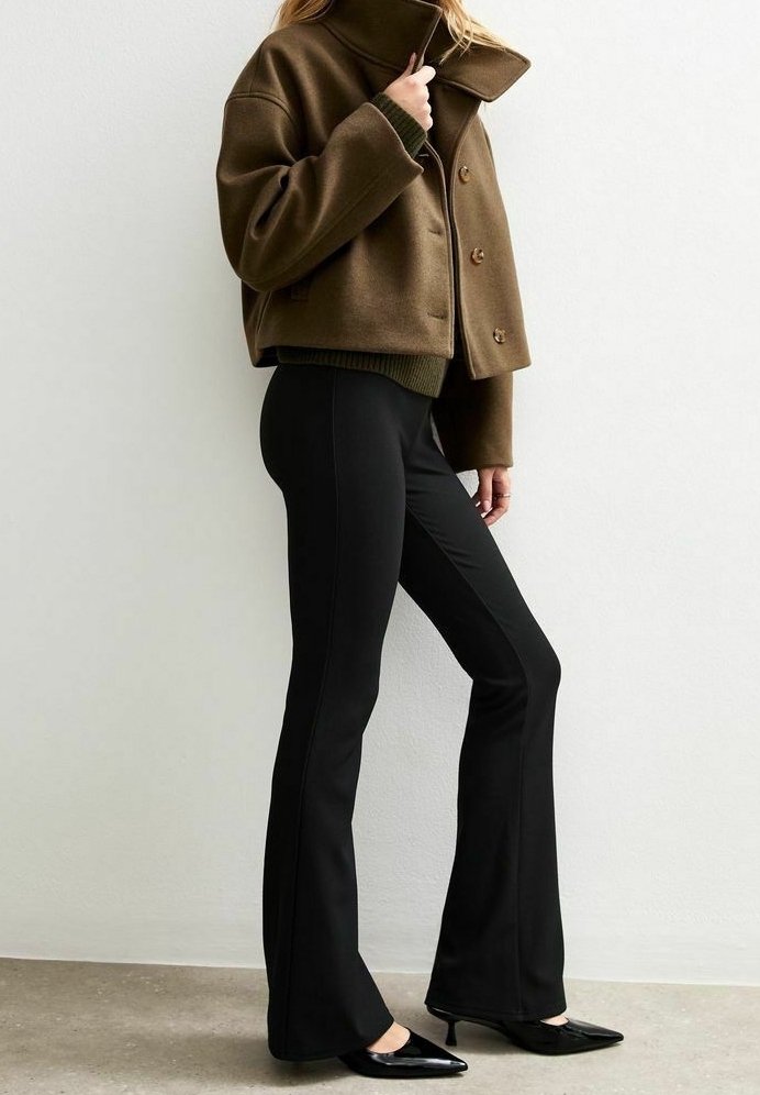 Femme portant une veste courte vert olive, un pull vert foncé, un pantalon flare noir ajusté et des chaussures noires pointues à petit talon, debout de côté.