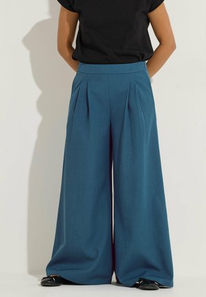 Persona con pantalones azules de talle alto y pierna ancha con pliegues delanteros y zapatos negros, de pie sobre un fondo claro.