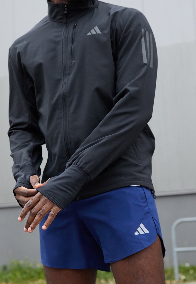 Personne portant une veste noire à manches longues zippée et un short de sport bleu avec un logo blanc, ajustant sa manche en extérieur devant un mur gris.