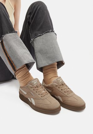 Person siddende iført optrukne mørke jeans, beige sokker og beige Reebok sneakers på hvid baggrund.
