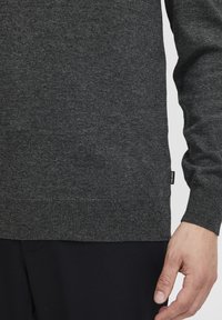 Maglione a maglia grigio scuro con orlo e polsini a coste, realizzato in un materiale morbido. Include un piccolo logo su un lato.