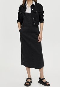 Černé džíny celkově šaty se rovnými liniemi, předními kapsami a detailním zapínáním na boku. Kombinováno s krátkou černou džínovou bundou a plochými sandály.