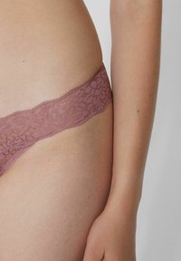 Koronkowe stringi w stonowanym mauve, z floralnym wzorem na pasku, miękką teksturą i niskim stanem, podkreślające okolice bioder.