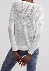 Weißer Strickpullover mit lockerer Passform, breitem Ausschnitt und langen Ärmeln. Strukturmuster mit offener Webart, kombiniert mit blauen Jeans.