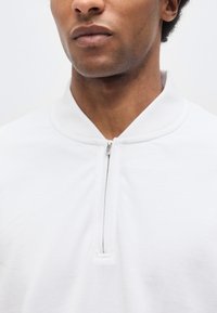 Κοντινό πλάνο ενός ατόμου που φοράει λευκό τοπ με φερμουάρ, με το φερμουάρ να είναι μερικώς κλειστό στον γιακά.