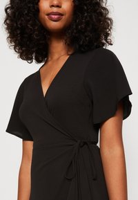 Robe noire à encolure en V avec des manches courtes volantées, en tissu texturé, et un détail à nouer à la taille. Silhouette lisse et fluide.