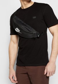 Muž v černém tričku a hnědých kalhotách s černou ledvinkou Nike připevněnou přes hruď.