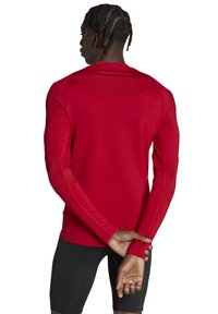 Rotes Longsleeve-Sportoberteil aus feuchtigkeitsableitendem Material, mit enganliegendem Design und flachen Nähten mit kontrastierender oranger Naht.