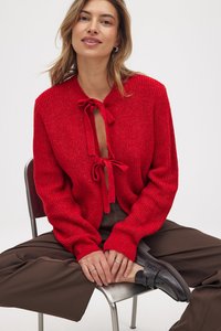 Rote Strickjacke mit einer Bindegürteldetail, langen Ärmeln, gerippter Textur, getragen über braunen Hosen, kombiniert mit schwarzen Slippern.