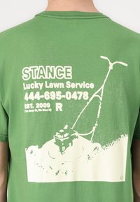 T-shirt de algodão verde com um gráfico branco de uma cortadora de relva, detalhes do negócio e idade. Gola redonda com mangas curtas.