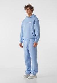 Šviesiai mėlynas džemperis su gobtuvu ir suderintos kelnes, pagamintos iš minkštos medžiagos, su priekiniu kišeniu ir maža grafika ant krūtinės. Balti sportbačiai.