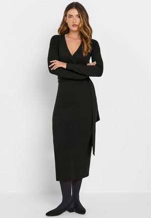 Black long-sleeve wrap dress made of soft fabric, featuring a deep V-neckline and a side tie, paired with black tights and flat shoes.