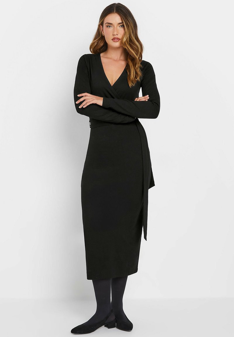 Schwarzes langärmeliges Wickelkleid aus weichem Stoff, mit einem tiefen V-Ausschnitt und einer seitlichen Schleife, kombiniert mit schwarzen Strumpfhosen und flachen Schuhen.